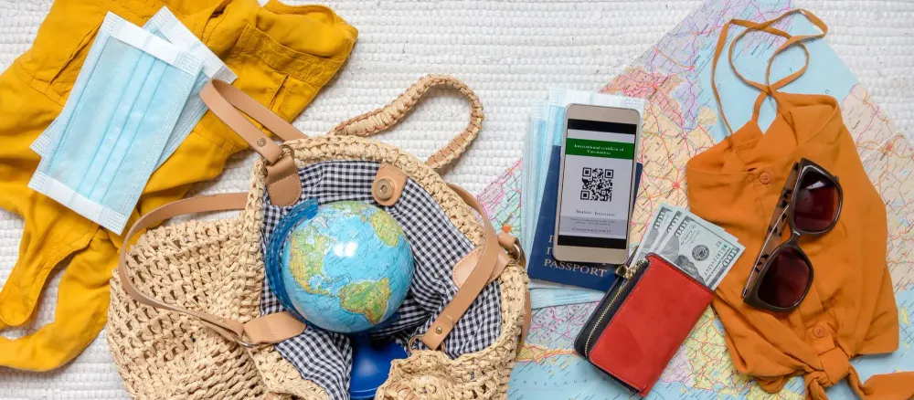 A travel themed flat lay featuring a small globe, face masks, a COVID-19 immunity passport, sunglasses, cash, and a passport